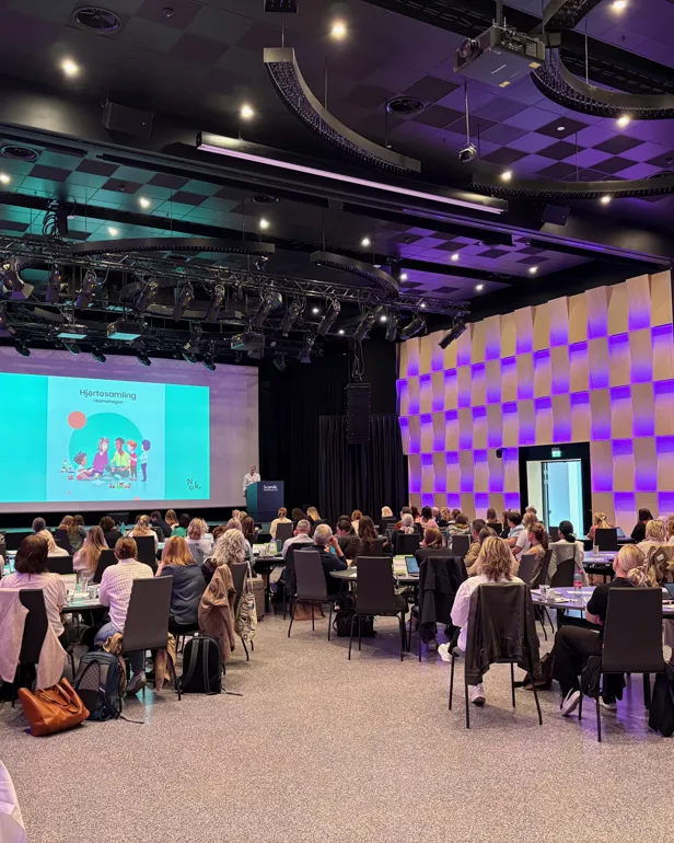 Ei gruppe menneske som sit i eit rom med ein stor skjerm