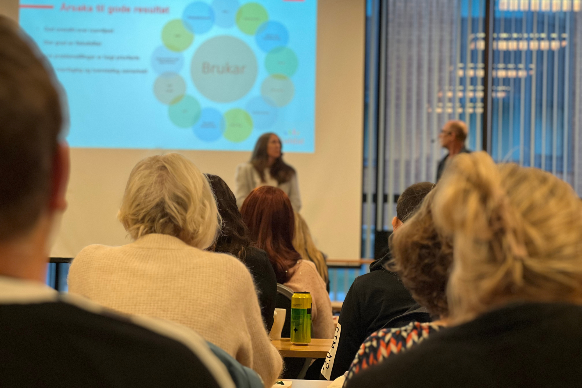 Ei gruppe menneske som sit i eit rom og ser på eit par som underviser